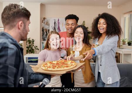 Prendi una fetta di pizza. Amici eccitati e multicolturati hanno ottenuto pizza appena sfornata mentre hanno partito del karaoke a casa Foto Stock