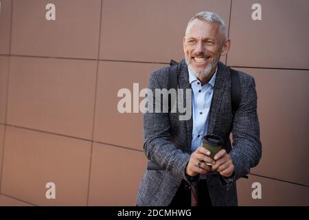 Felice uomo d'affari moderno di mezza età sorridente mentre si alza in piedi all'aperto con scooter elettrico e una tazza di caffè Foto Stock