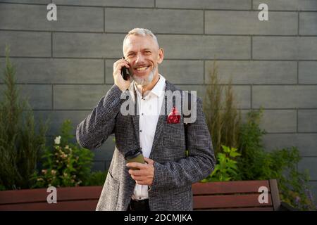 Su chiamata. Ritratto di allegro uomo d'affari di mezza età in elegante tuta sorridente mentre si parla al telefono, in piedi all'aperto con una tazza di caffè Foto Stock