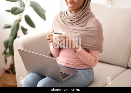 Lavoro on line, istruzione, comunicazione. Donna nera irriconoscibile in hijab che usa il laptop e beve il caffè a casa Foto Stock