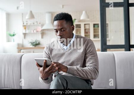 Uomo africano serio che usa le app per tablet digitali seduti sul divano a casa. Foto Stock