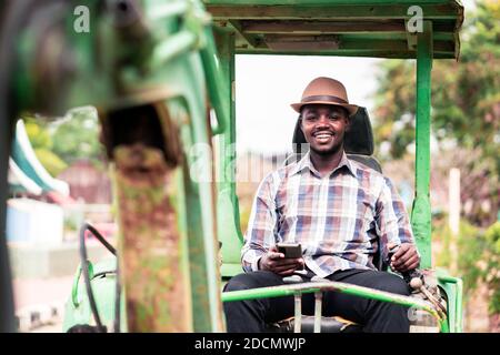 Lavoratore africano che guida attrezzature pesanti per l'edilizia retroescavatore con l'uso di smartphone Foto Stock