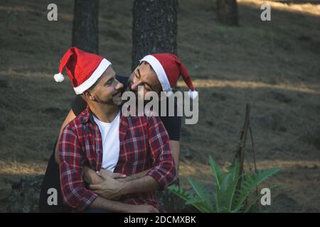 Scena romantica di giovane coppia gay con cappelli rossi di Natale nel parco forestale. Uomini sorridenti che si abbracciano nella stagione invernale all'aperto in natura Foto Stock