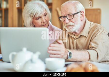 Donna anziana che guarda il marito mentre lavora con il computer portatile a casa. Concetto di famiglia e di famiglia Foto Stock