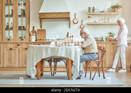 Sorridente uomo anziano che scrive sul computer portatile mentre sua moglie prepara la colazione vicino a lui. Concetto di stile di vita domestico Foto Stock