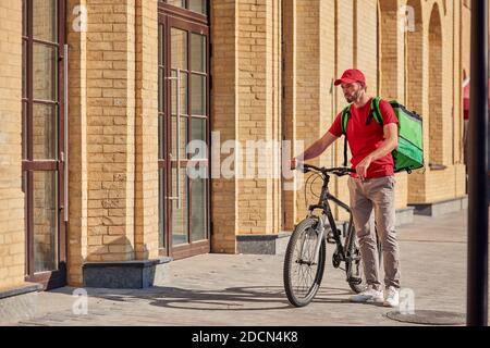 Consegna del cibo in bicicletta. Corriere maschile giovane in uniforme rossa con zaino termico in piedi sulla soleggiata strada della città Foto Stock