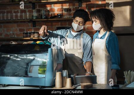 Giovane afroamericano ragazzo in grembiule e maschera protettiva insegna donna apprendista a lavorare con la macchina espresso Foto Stock