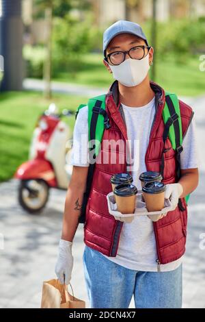 Il corriere asiatico giovane indossa maschera protettiva per il viso e guanti che tengono quattro tazze di caffè e sacchetto di carta con cibo, guardando la macchina fotografica mentre si trova in piedi per strada. Consegna sicura durante la pandemia di covid 19 Foto Stock