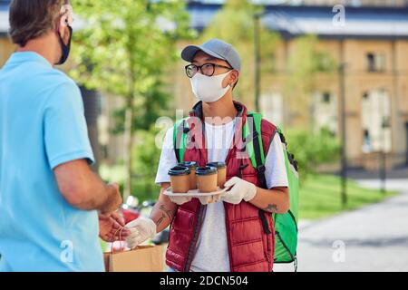 Concetto di soggiorno a casa. Giovane corriere asiatico in maschera protettiva e guanti che consegna quattro tazze di caffè al cliente mentre si trova in una strada soleggiata. Consegna di cibo durante l'epidemia di covid 19 Foto Stock