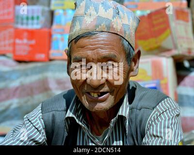 L'anziano trader nepalese Tamang indossa un cappello tradizionale nepalese (dhaka topi) e sorride per la telecamera di fronte alla sua merce al mercato settimanale. Foto Stock