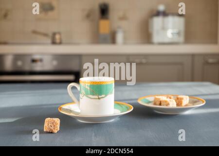 Una piccola e bella tazza di caffè si trova su una superficie grigia scura su uno sfondo sfocato dell'interno di una cucina accogliente in una mattina o giorno di sole. Accanto a. Foto Stock