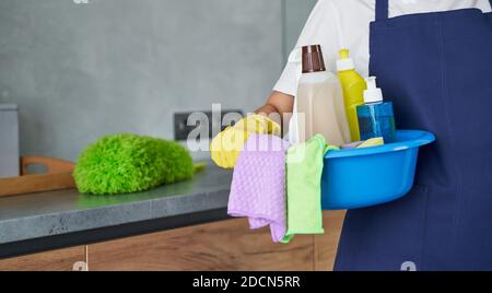 Primo piano di pulizia signora che tiene contenitore pieno di prodotti di pulizia e attrezzature mentre in piedi nella cucina moderna. Lavori di pulizia e pulizia delle camere, concetto di servizio di pulizia Foto Stock