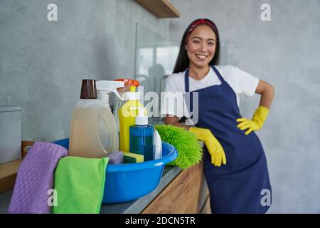 Primo piano di contenitore pieno di prodotti per la pulizia e attrezzature con donna giovane, signora pulizia in piedi sullo sfondo. Lavori di pulizia e pulizia delle camere, concetto di servizio di pulizia Foto Stock