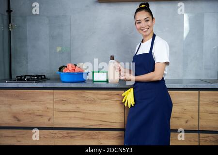 I migliori prodotti. Giovane donna allegra, donna che si pulisce sorridendo alla macchina fotografica, in piedi con detersivo liquido per bucato mentre si caricano i vestiti sporchi nella lavatrice. Concetto di servizio di pulizia professionale della casa Foto Stock