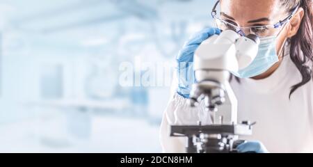 Tecnico di laboratorio femminile in occhiali protettivi, guanti e maschera facciale si siede accanto a un microscopio in laboratorio. Foto Stock