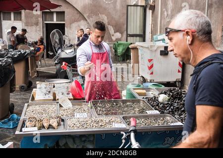 CATANIA, SICILY, ITALY - Oct 15, 2016: Il venditore di frutti di mare che sgocciolano l'acqua sui molluschi per mantenerli freschi, il cliente potenziale che cerca sopra. Contrassegno Catania Foto Stock