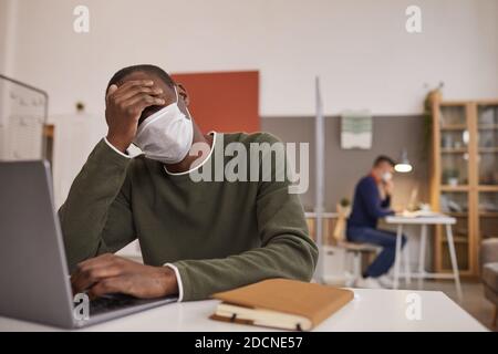 Ritratto di un uomo afro-americano stanco e malato che indossa una maschera e utilizza un computer portatile mentre lavora alla scrivania in ufficio, spazio per la copia Foto Stock