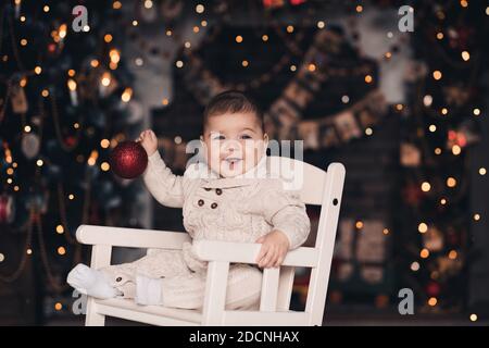 Sorridente bambino felice sotto 1 anno che tiene la sfera di Natale seduta nella sedia della roccia sopra l'albero con le luci incandescenti vicino in su. Guardando la fotocamera. Inverno Foto Stock