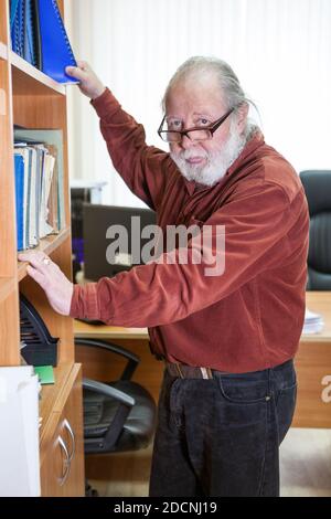 L'uomo anziano bearded mette i documenti in una mensola in ufficio. Camicia marrone e occhiali Foto Stock