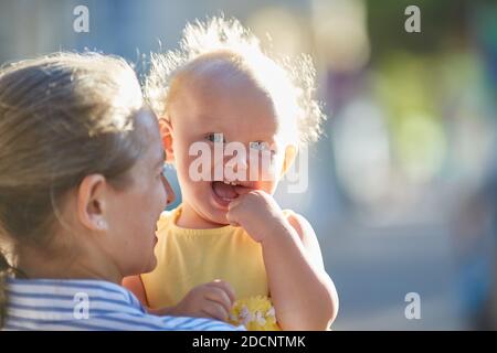 Felice bambino sorridente tra le braccia della madre. Bella raggi di sole sulla strada in una giornata estiva. Foto Stock