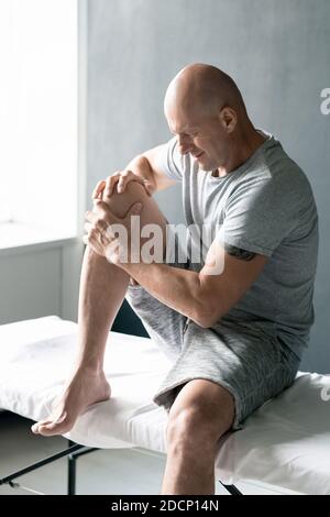 Sportivo maturo e calvo contemporaneo con maglietta grigia e pantaloncini massaggianti ginocchio Foto Stock