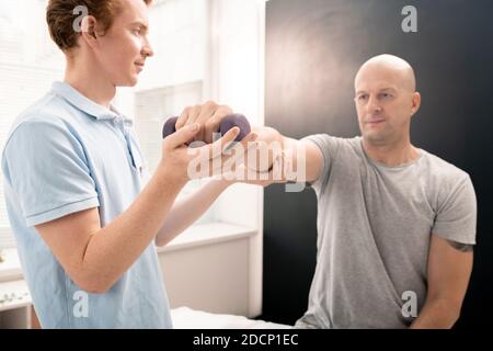 Giovane fisioterapista che aiuta il paziente a sollevare e allungare il braccio con dumbbell Foto Stock