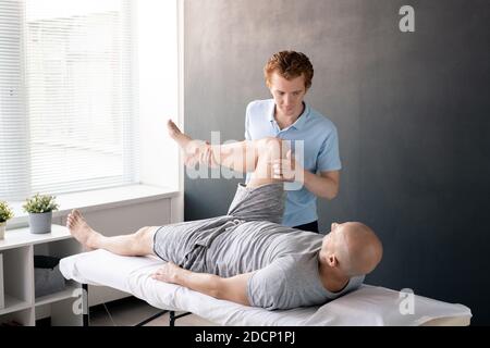 Fisioterapista maschile serio che guarda il paziente con la gamba piegata nel ginocchio Foto Stock