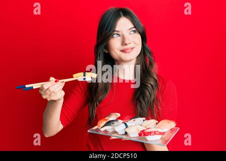 Giovane bella ragazza caucasica mangiare sushi utilizzando chopsticks sorridendo guardando al lato e fissando via pensare. Foto Stock