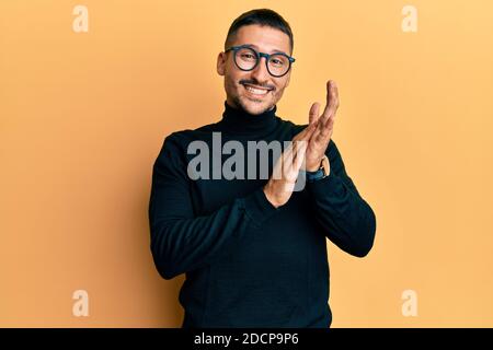 Bell'uomo con tatuaggi che indossano felpa con dolcevita e occhiali che si aggrappano e applaudono felici e gioiosi, sorridendo insieme mani orgogliose Foto Stock