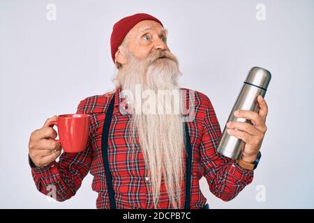 Vecchio uomo anziano con capelli grigi e barba lunga che tiene una tazza di caffè e termo sorridente guardando al lato e fissando via pensare. Foto Stock