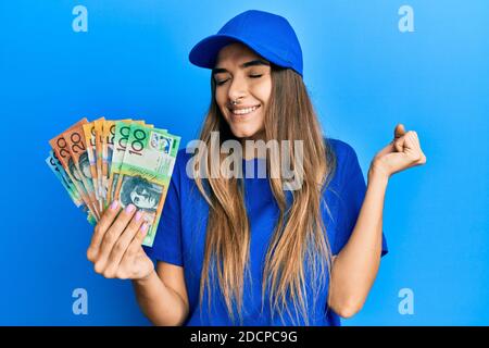 Giovane donna brunetta con piercing che indossa uniforme di consegna tenendo australiano i dollari celebrano il successo con il sorriso felice e l'espressione del vincitore Foto Stock