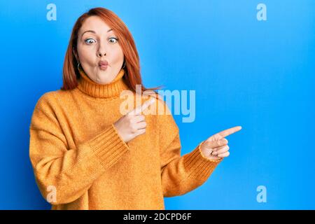 Bella donna rossa che punta le dita verso il lato facendo faccia di pesce con la bocca e squinting occhi, pazzo e comico. Foto Stock