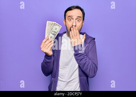 Uomo bello di mezza età che tiene mazzo di banconote di dollari su sfondo viola isolato che copre la bocca con la mano, scioccato e impaurito per errore. S Foto Stock