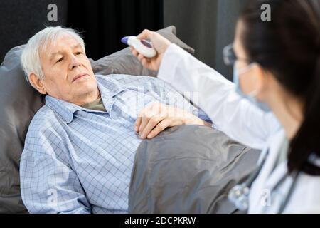 Ammalato anziano che guarda una giovane dottoressa senza contatto termometro Foto Stock