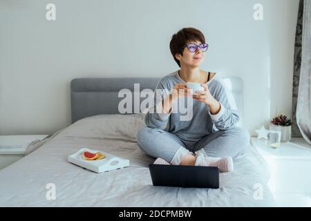 Donna caucasica in occhiali, vestiti casual a casa bevendo il suo caffè di prima colazione durante il lavoro sul computer portatile sul suo letto. Lavoro remoto da casa. Ora a. Foto Stock