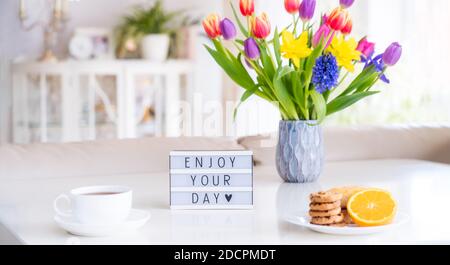 Concetto di Buongiorno. Colazione romantica - fiori freschi, tazza di bevanda calda, biscotti, arancio, scatola luminosa con messaggio Godetevi la vostra giornata sul tavolo in marmo con Foto Stock