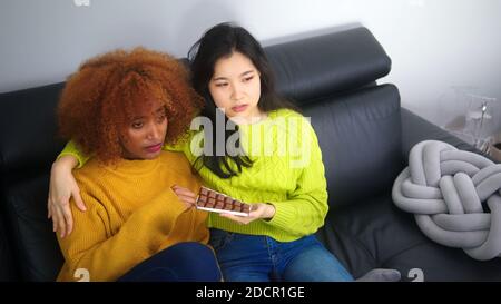 Donna triste con cioccolato e allegra fidanzata seduta sul divano. Foto di alta qualità Foto Stock