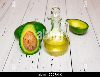 Olio di avocado in una caraffa di vetro, avocado accanto ad esso. Vista laterale, sfondo bianco in legno. Cibo vegetariano sano, vitamine. Foto Stock