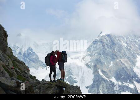 Vista laterale di coppie amanti arrampicatori con zaini baciando, bellissimo paesaggio di montagne sullo sfondo. Trekking, turisti che raggiungono la vetta insieme. Natura selvaggia con vista mozzafiato. Turismo sportivo nelle Alpi. Foto Stock