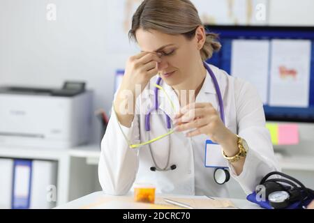 Stanco medico che si siede a tavola nel suo ufficio e guarda dopo gli occhi Foto Stock