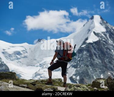 Zinal, Svizzera - 19 luglio 2019: Uomo viaggiatore con zaino che tiene una bottiglia d'acqua e godere della vista delle colline innevate sotto il cielo blu durante l'escursione in montagna. Concetto di viaggio, alpinismo. Foto Stock