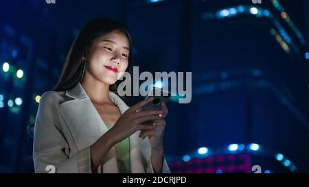 Ritratto di sorridente bella giovane donna asiatica utilizzando smartphone in la città di notte Foto Stock