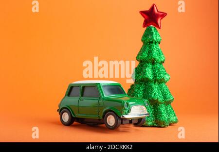 Albero di Natale di Capodanno con stella rossa sulla parte superiore vicino giocattolo auto verde . Sfondo arancione. Albero e auto in miniatura creativa di Natale. Concetto di consegna dei regali Foto Stock