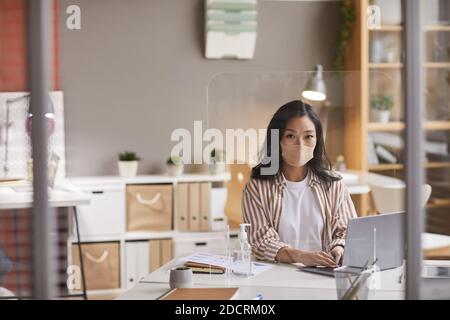 Ritratto della giovane donna asiatica che indossa la maschera facciale e utilizza il computer portatile mentre si siede alla scrivania in ufficio, spazio per la copia Foto Stock