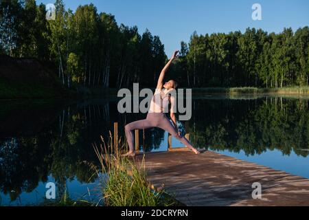 Giovane donna presso il lago che pratica yoga si muove su piattaforma di legno. Bella giovane donna che si esercita in natura, sano stile di vita giovani positivi Foto Stock