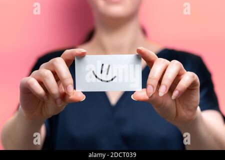 Una donna blurry tiene una carta bianca con un emoticon sorridente disegnato su di essa. Primo piano. Sfondo rosa Foto Stock