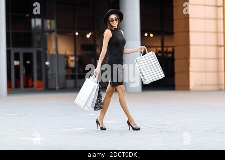 Shopping viaggio, felice attraente ragazza in un elegante vestito nero, cappello e occhiali da sole, con borse per lo shopping, sorride mentre cammina lungo la strada. Shopping Foto Stock