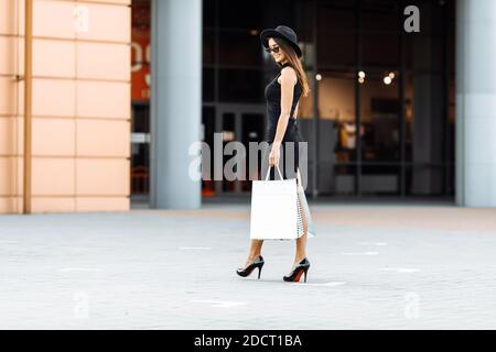 Shopping viaggio. Black Friday. Una ragazza attraente felice in un abito nero elegante, cappello e occhiali da sole, con borse per lo shopping, sorride mentre cammina lungo la s. Foto Stock