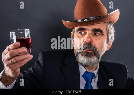 L'uomo anziano in tuta con cravatta e cappello da cowboy valuta la trasparenza del vino Foto Stock