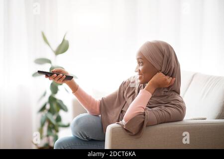 Vista laterale della bella donna nera in hijab tradizionale con telecomando e guardare la TV all'interno, vista laterale Foto Stock